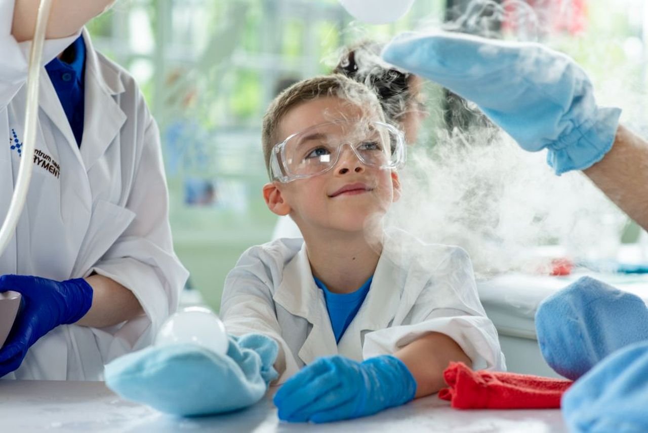 Chłopiec w fartuchu, rękawicach i goglach ochronnych obserwuje doświadczenie chemiczne.