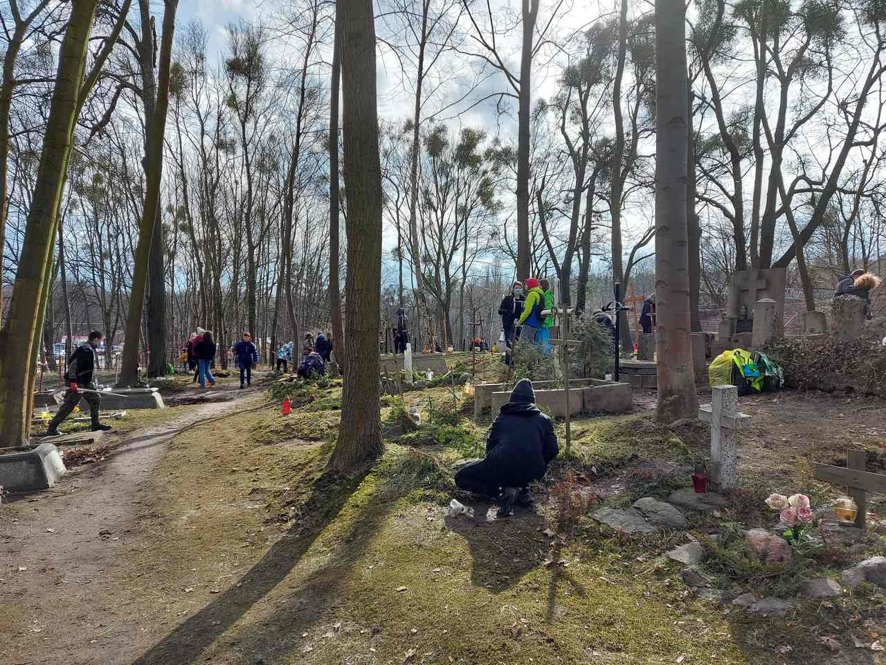 Grupa osób sprzątająca zaniedbaną nekropolię. Na około stare drzewa, zieleń, słońce prześwietla przez gałęzie drzew.