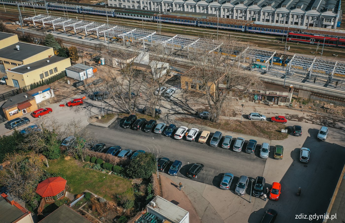 widok z góry na teren parkingu przy stacji SKM, widoczne zaparkowane pojazdy, tory kolejowe, budynki