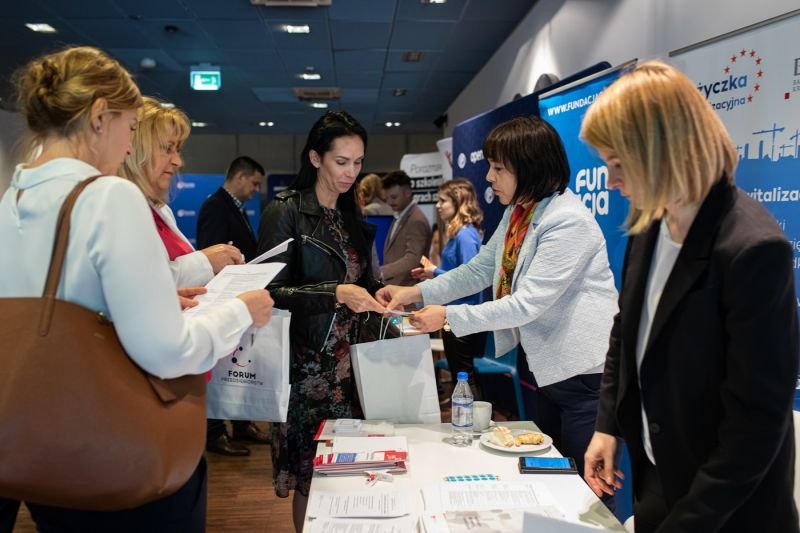 Hol PPNT Gdynia. Grupa osób ogląda drukowane materiały promocyjne leżące na stole. Osoby stoją po obu stronach stołu