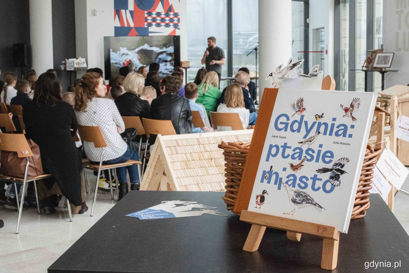 Foyer Muzeum Miasta Gdyni. W tle tłum na widowni i prezentacja na ekranie, na pierwszym planie stolik ze sztalugą, na sztaludze książka 
