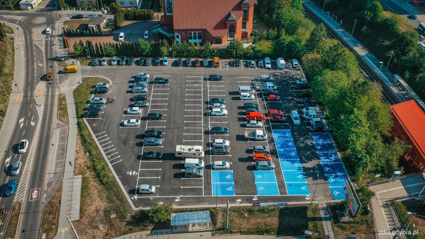 widoczny z góry parking, a na nim zaparkowane pojazdy. Na parkingu widoczne oznakowanie, linie oraz niebieskie miejsca postojowe dla osób z niepełnosprawnościami. W tle jezdnia, ruch uliczny i drzewa.