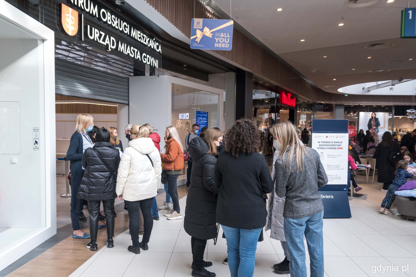 Wnętrze Centrum Riviera. Widoczne Centrum Obsługi Mieszkańca Urzędu Miasta Gdyni. Ludzie przed wejściem, rozmawiają w małych grupach