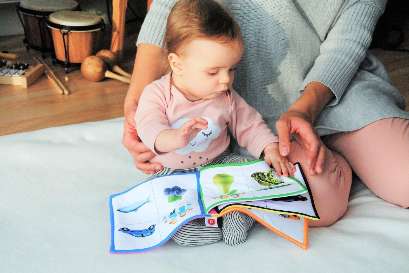 Na podłodzie na dywaniku małe dziecko, obok jej mama widoczna jedynie do połowy. Razem czytają, oglądają książeczkę dla dzieci.