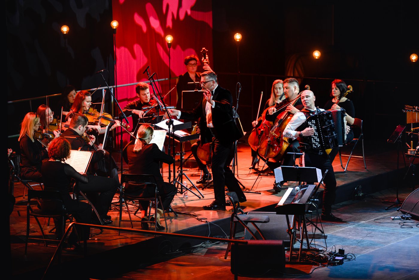 Oświetlona czerwonym kolorem scena. Na niej zespół muzyczny - osoby grające na instrumentach smyczkowych oraz dyrygent, a także mężczyzna grający na akordeonie.