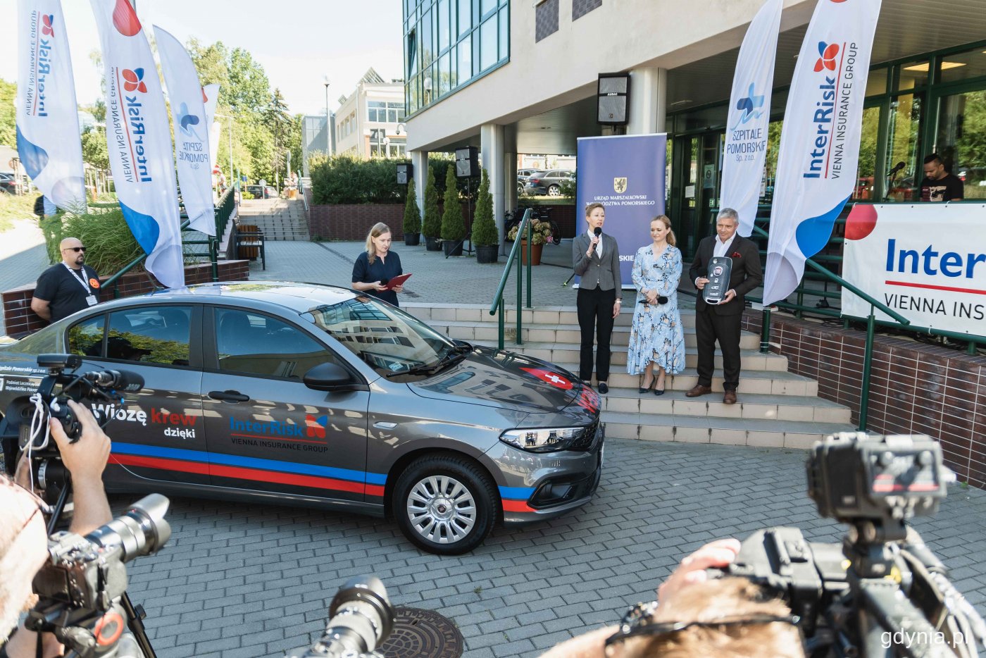 Plac przed wejściem do Szpitala w Redłowie. Stoi zaparkowany oznaczony samochód, obok na schodach kilka osób w formalnych strojach. Na dole kamery, aparaty i dziennikarze.