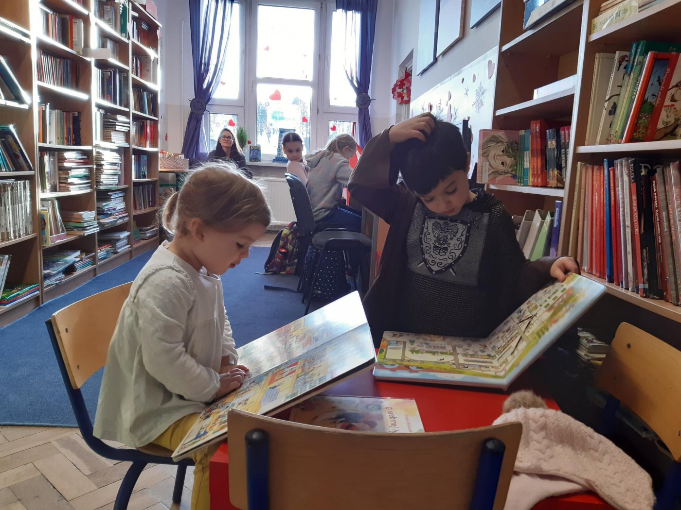 Dzieci czytają książki przy stolikach w bibliotece.