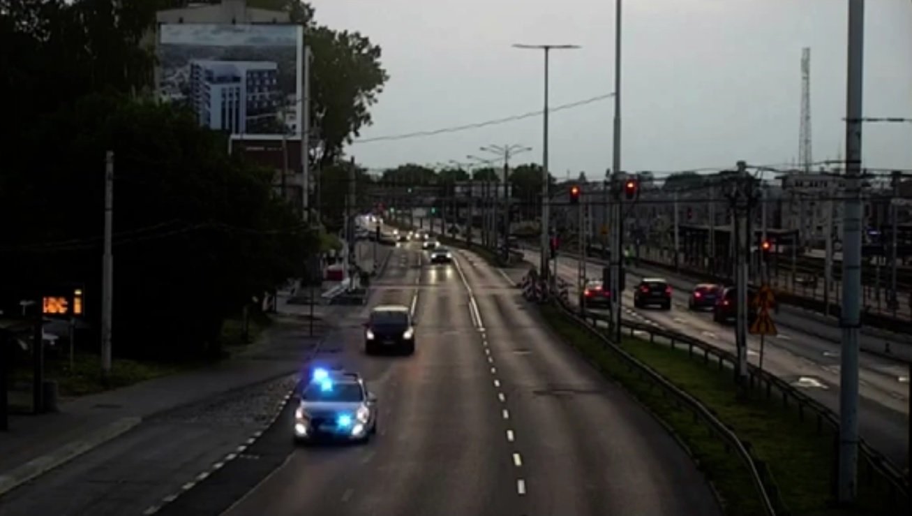 Stop-klatka z monitoringu miejskiego. Po ulicy jedzie radiowóz na sygnale, za nim auto osobowe.