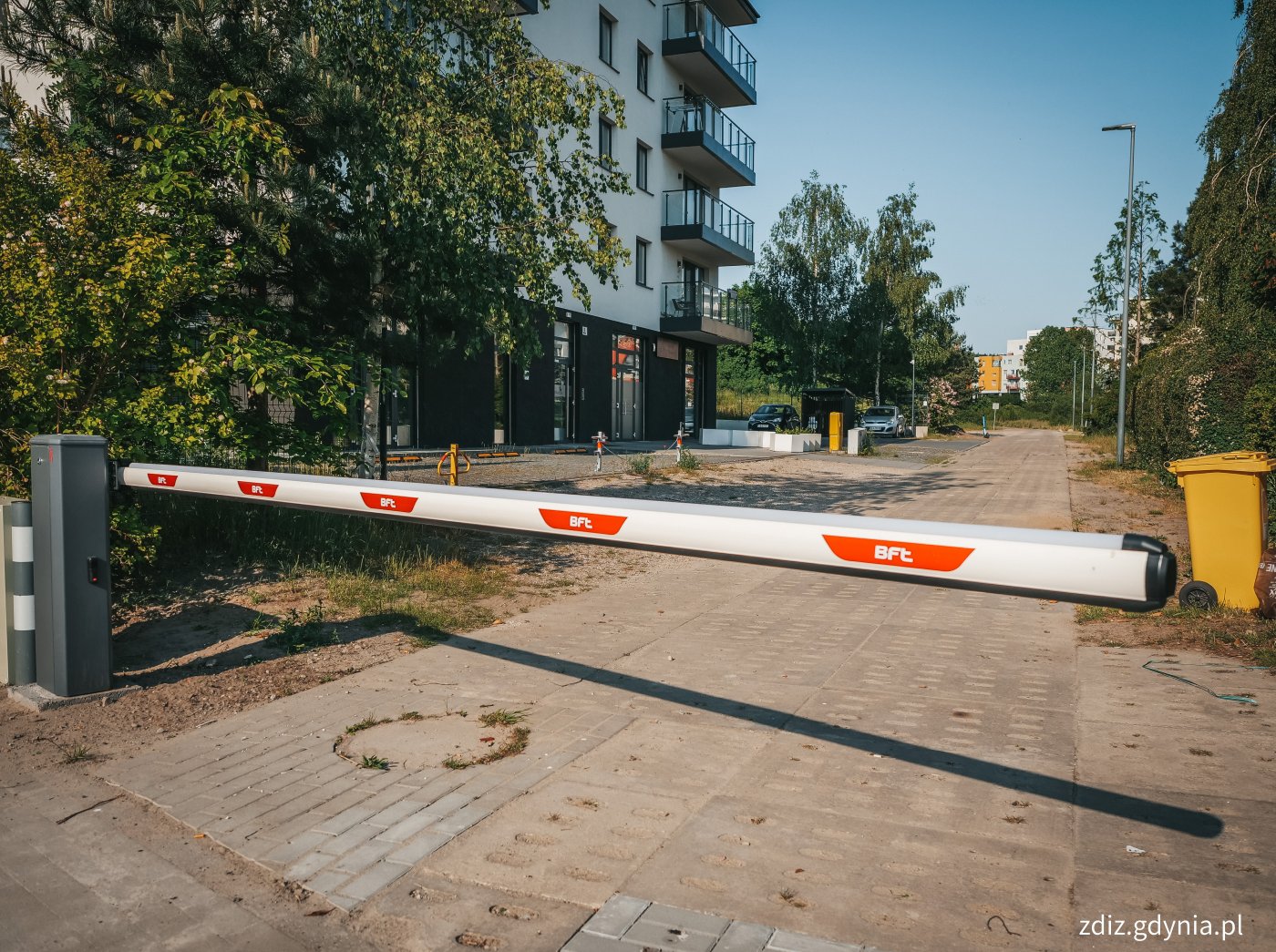 szlaban na drodze, widoczna nawierzchnia z płyt, w tle zieleń oraz budynki, szlaban koloru biało-czerwonego