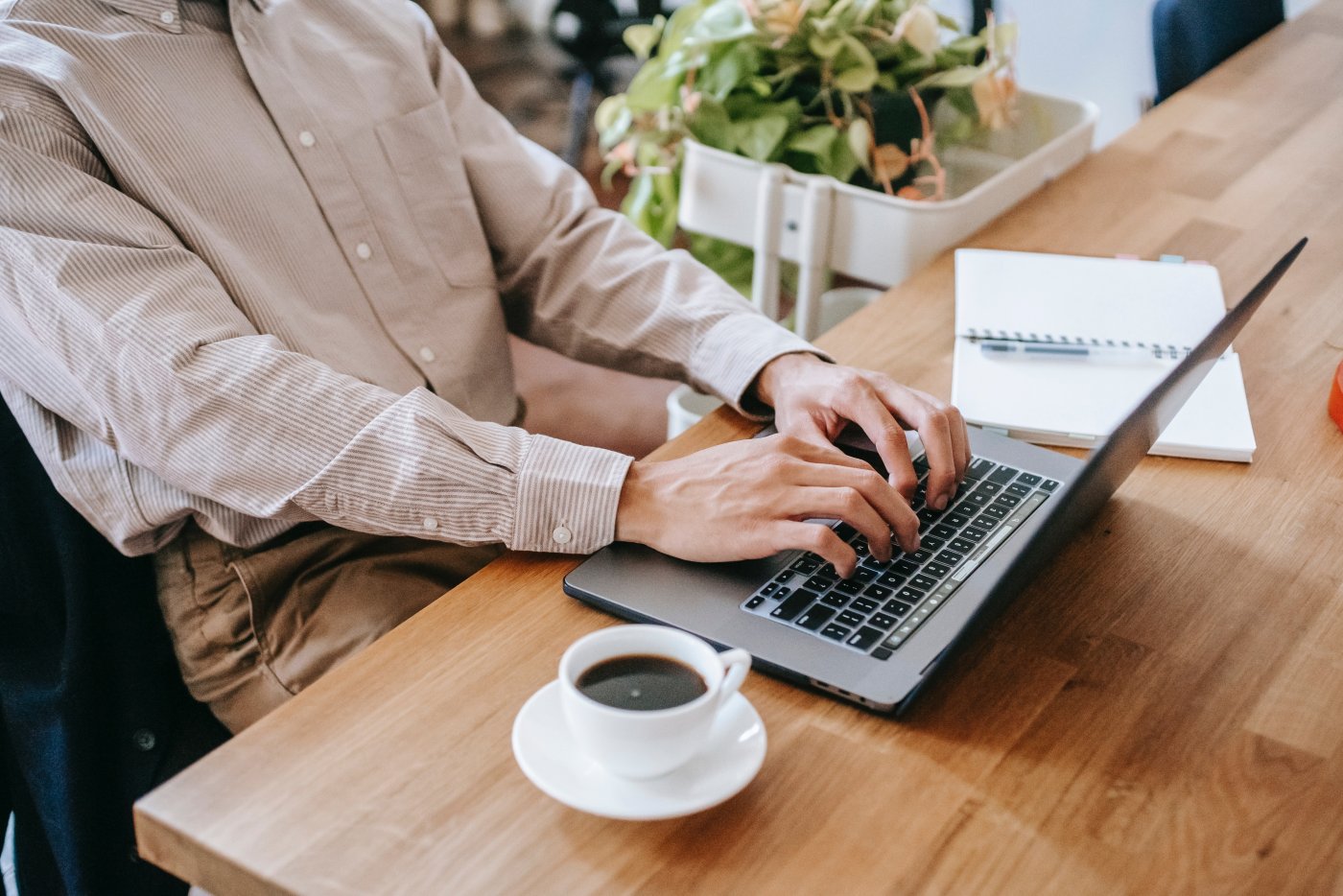 Mężczyzna siedzący przy biurku, pisze na laptopie. Obok laptopa ma filiżankę z kawą i notatnik.