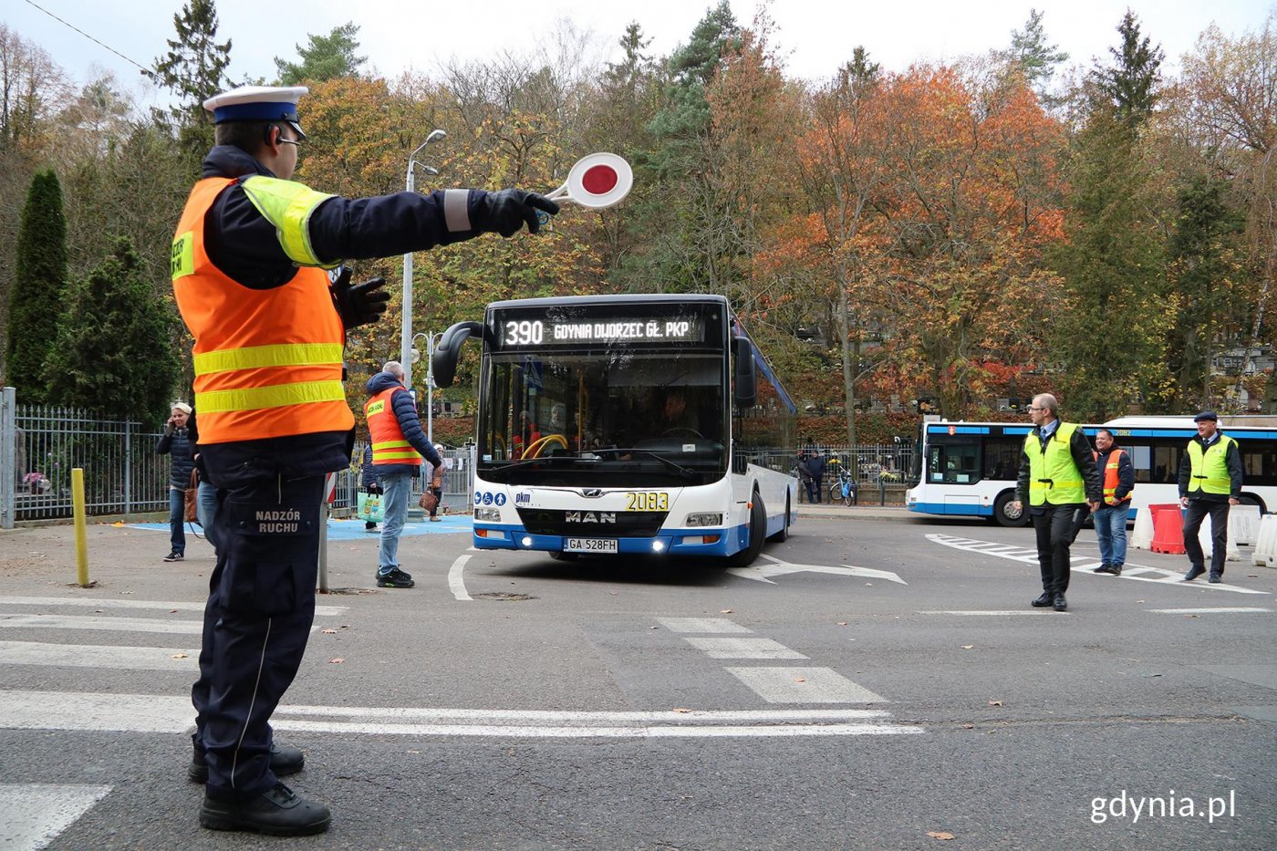 Funkcjonariusz wskazujący drogę dla autobusu na parkingu przy cmentarzu.