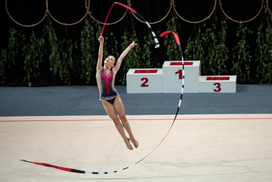Na zdjęciu dziewczyna w podskoku, w kolorowym kostiumie z wstęgą w ręcę podczas akrobacji gimnastycznych.