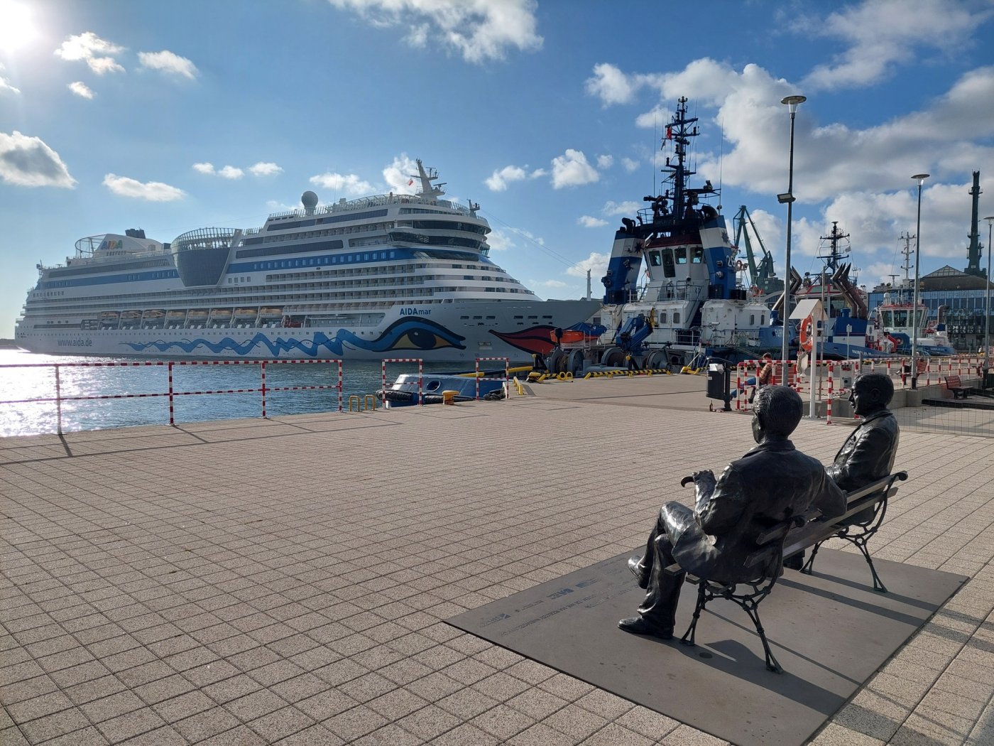 Ostroga Pilotowa, port Gdynia, nabrzeże, pomnik dwóch mężczyzn siedzących na ławce, w tle Muzeum Emigracji, statek wycieczkowy cumujący przy budynku, słoneczny dzień