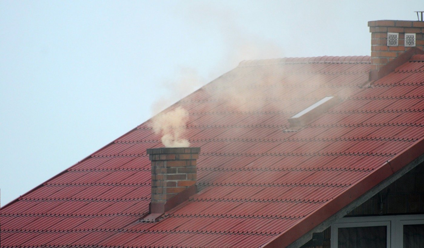 Dach z czerwonej dachówki z kominem, z którego wydobywa się szary, gęsty dym.