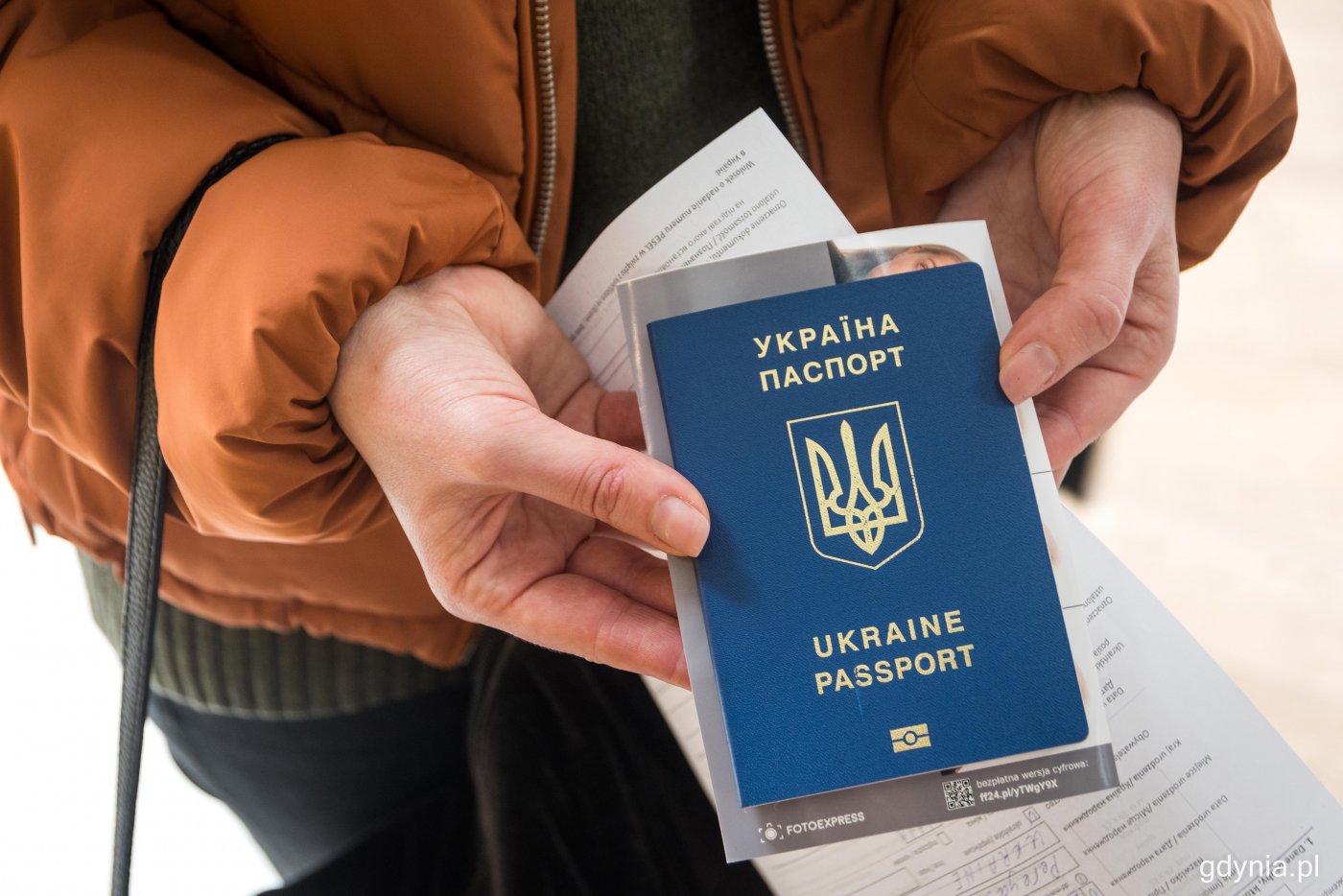 Kobiece dłonie, w dłoniach ukraiński, granatowy paszport, za okładką widoczny fragment zdjęcia twarzy.