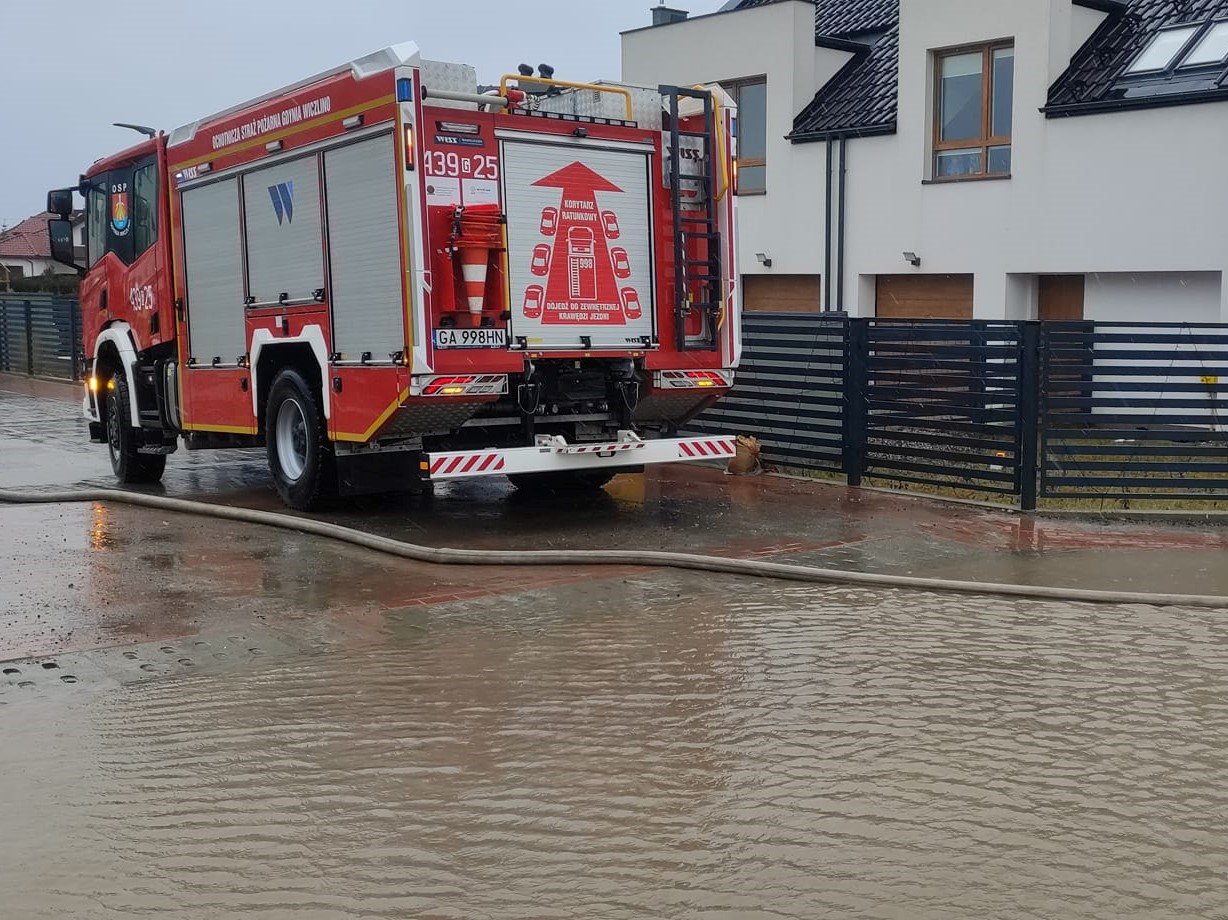 Deszczowa pogoda, ulica z kostki brukowej w Gdyni. Widoczny dom jednorodzinny, na chodniku stoi wóz straży pożarnej, ulica zalana wodą