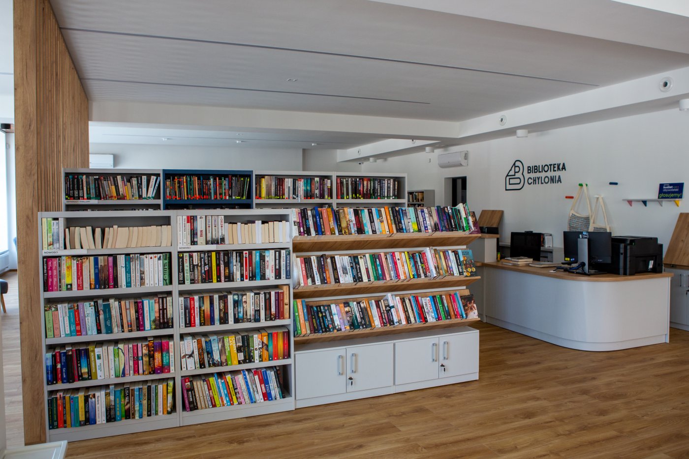 Na zdjęciu wnętrze fili Biblioteki Gdynia. Widoczne regały z książkami, po prawej stronie miejsce do obsługi czytelników. Podłogi i elementy mebli utrzymane w drewnie. Ściany koloru białego.