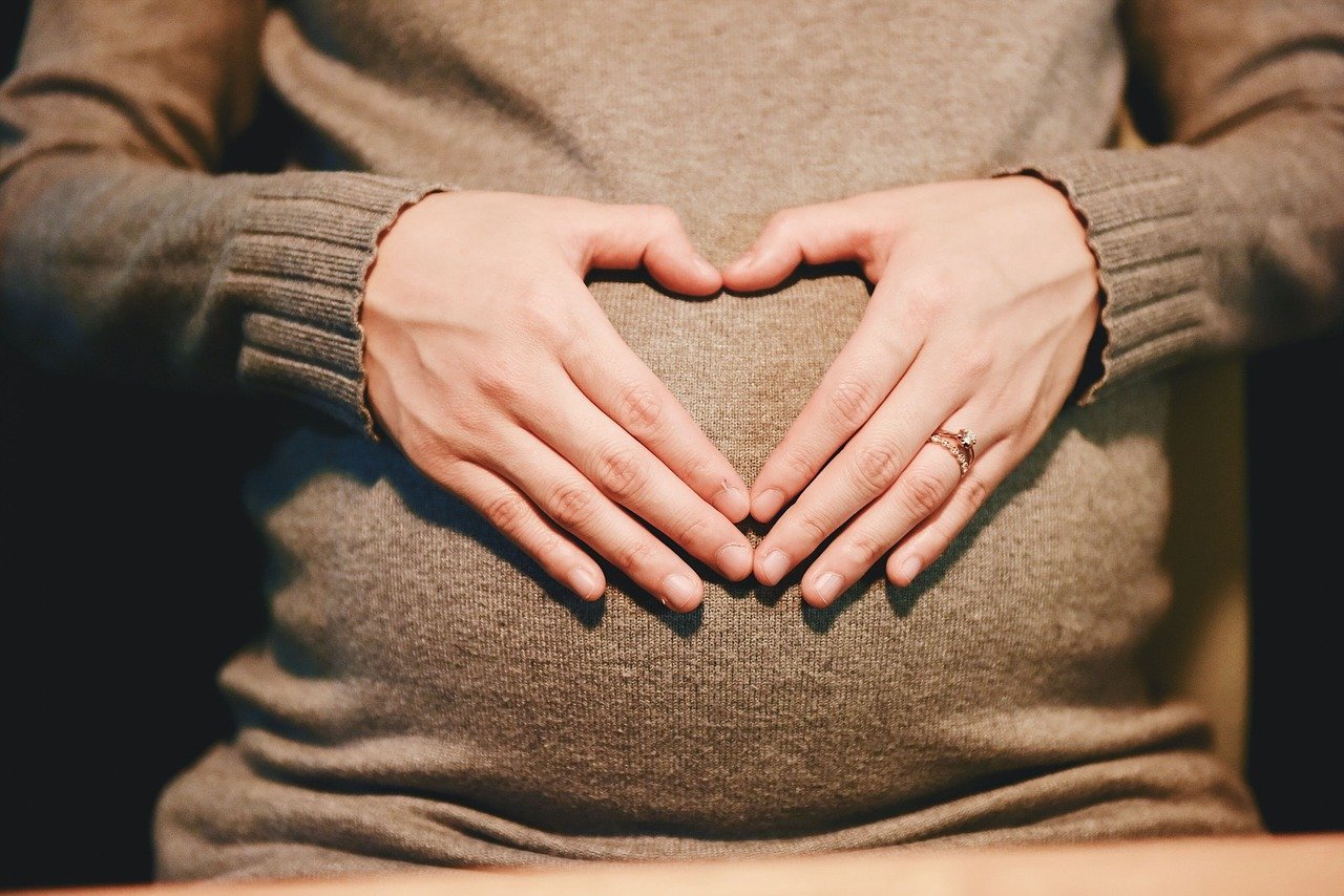Widoczny brzuch kobiety w ciąży, która na brzuchu z dłoni układa kształt serca