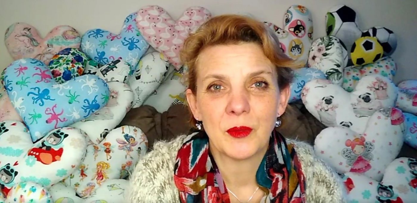 Kadr z filmu, na zdjęciu widoczna Justyna Sudzik, prezes fundacji Przytulny Domek, za panią prezes na opraciu kanapy dużo kolorowych ręcznie uszytych poduszek w kształcie serca.