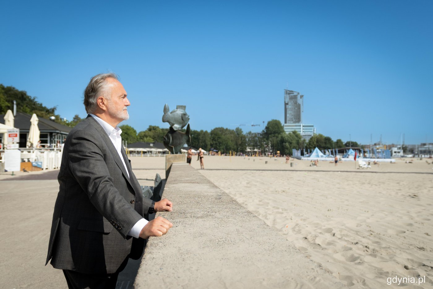 Wojciech Szczurek opiera się o betonowy falochron na bulwarze Nadmorskim w Gdyni. W tle plaża, budynki, ludzie na plaży, pomnik ryb.