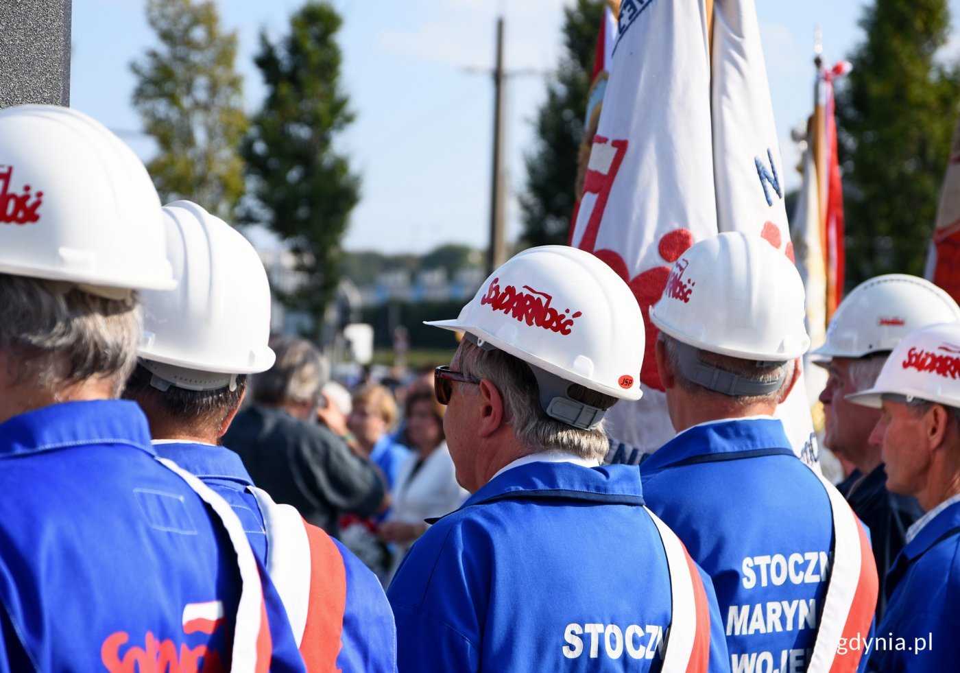 Mężczyźni stojący w szeregu, z białymi kaskami z napisem Solidarność i niebieskimi, roboczymi strojami. Ujęcie zza pleców.