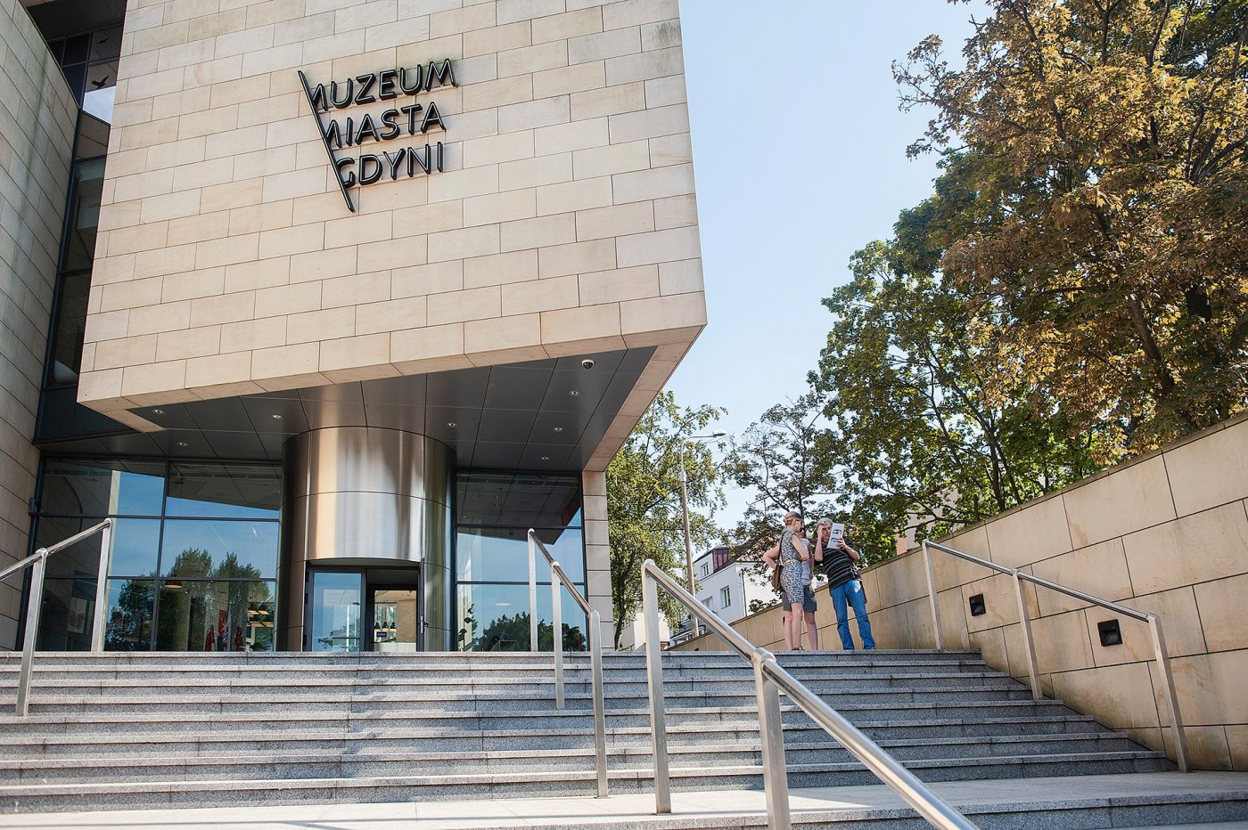 Muzeum Miasta Gdyni. Na pierwszym planie szare schody prowadzące do budynku. Powyżej widoczne wejście do muzeum oraz piaskowa bryła budowli. Na fasadzie widoczny napis-logotyp 