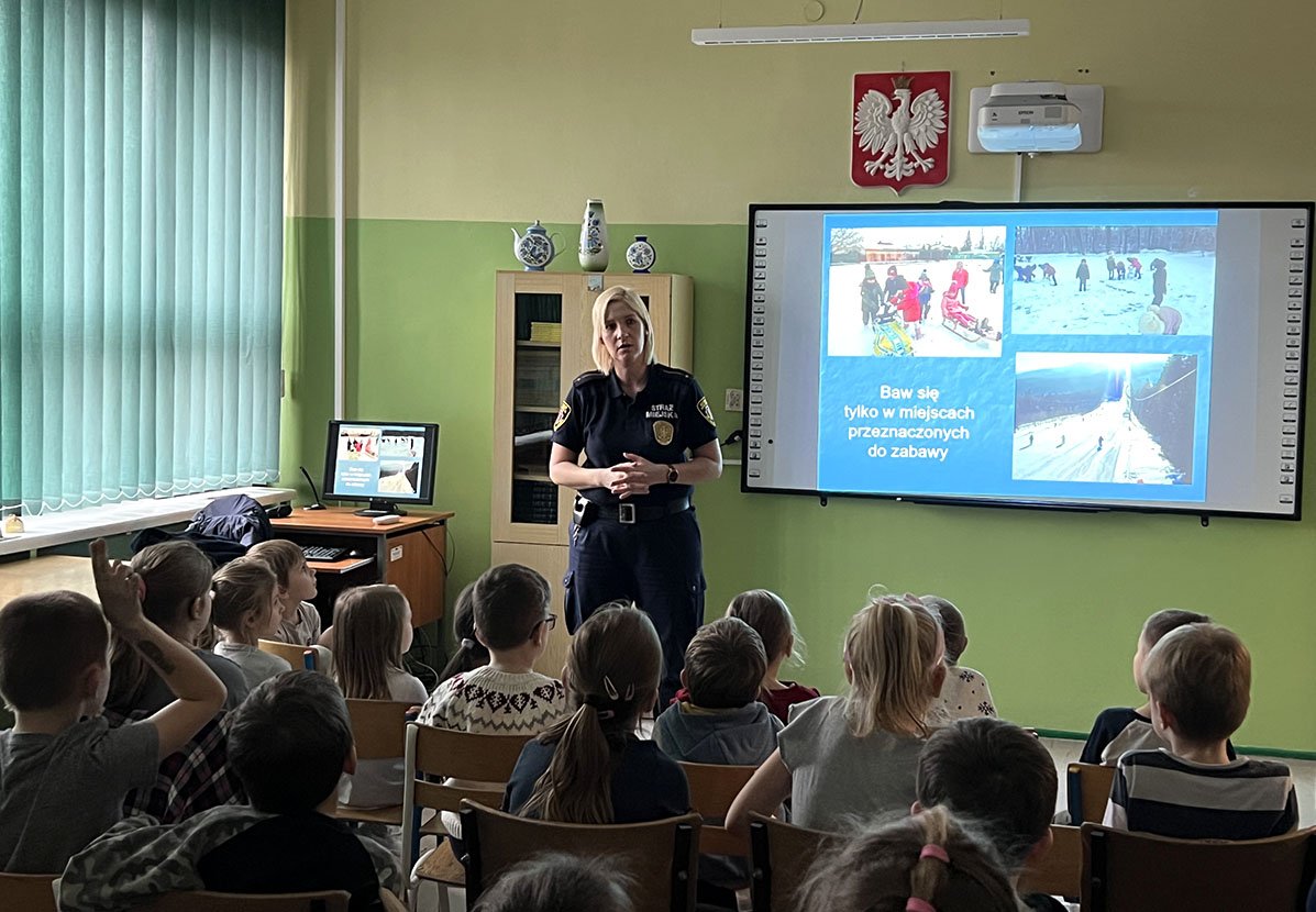 na zdjęciu strażniczka z jasnymi włosami stoi przed klasą z dziećmi w ławkach