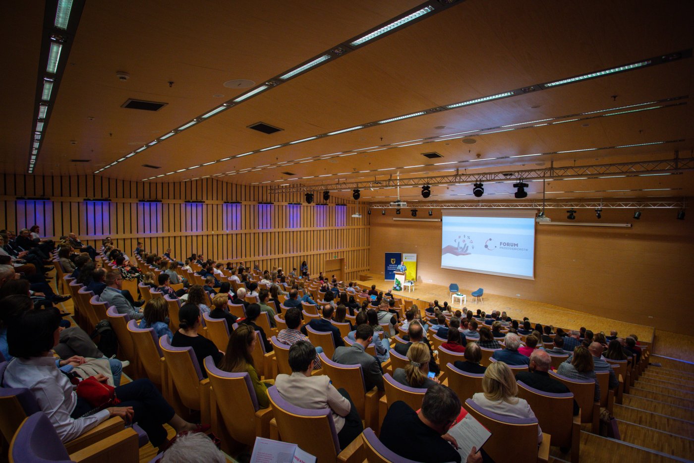 Sala konferencyjna w PPNT Gdynia, publicznośc na widowni, scena podświetlona, osoba przemawia zza pulpitu, wyświetlany obraz na ekranie