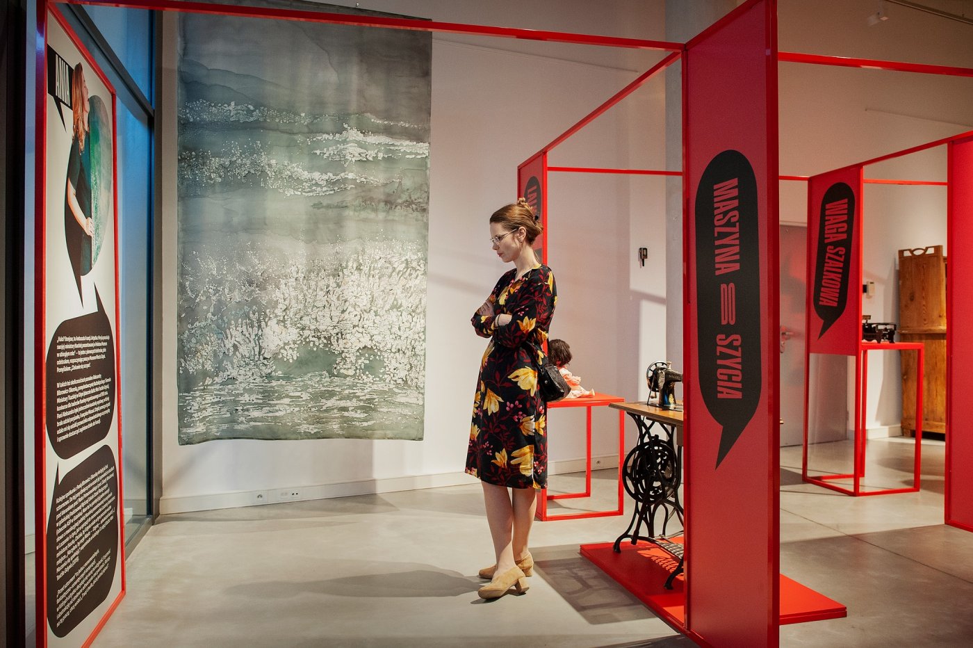 Kobieta w sukience, która ogląda wystawę „My o rzeczach \ rzeczy o nas” w Muzeum Miasta Gdyni. W tle stoją eksponaty: maszyna do szycia i lalka