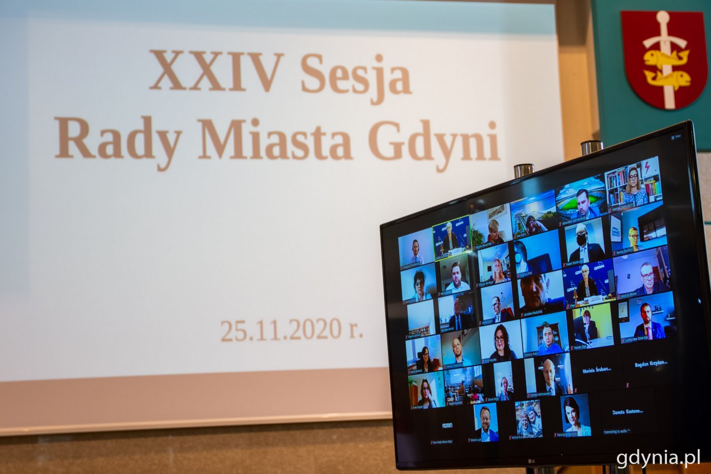 Po prawej stronie ekran LCD, na którym znajdują się kafelki z członkami Rady Miasta Gdyni, którzy połączeni są w ramach videokonferencji Zoom podczas XXIV sesji Rady Miasta Gdyni. W tle duży ekran z napisem XXIV sesja Rady Miasta Gdyni.