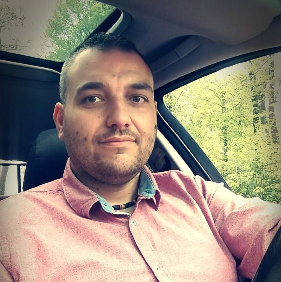 na zdjęciu młody ciemnowłosy mężczyzna w różowej koszuli za kierownicą