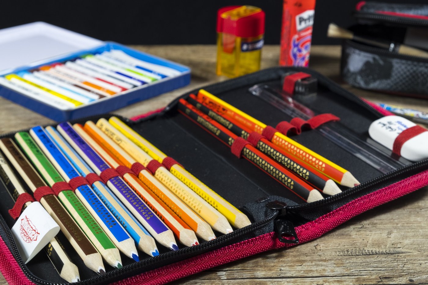 Piórnik z kolorowymi kredkami, linijką, ołówkami, temperówką i gumką do ścierania. W tle inne przybory szkolne // fot. pixabay.com