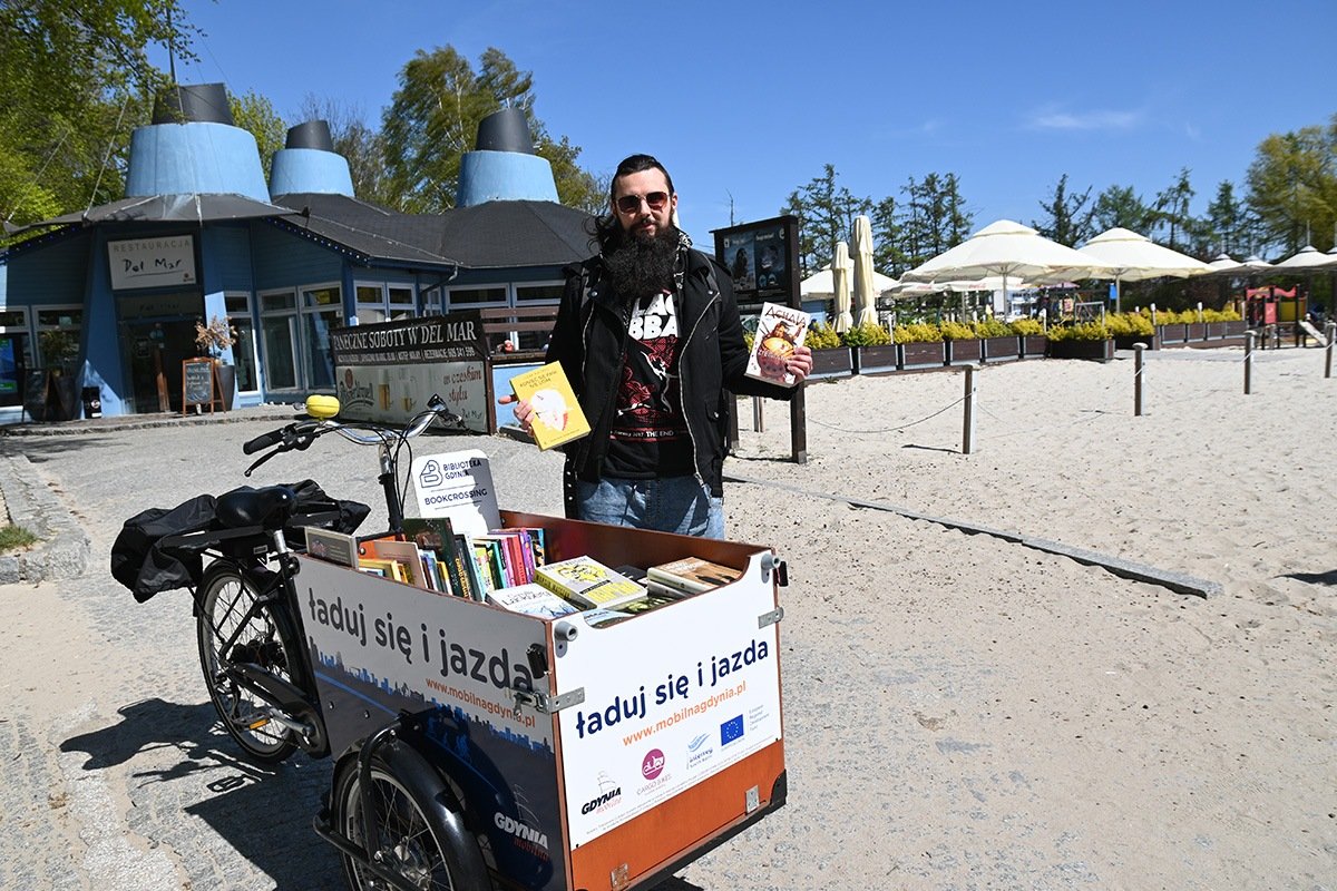 na zdjęciu rower cargo na plaży załadowany książkami, przy rowerze czarnowłosy uśmiechnięty mężczyzna