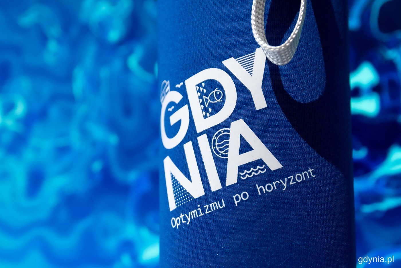 Widoczny fragment butelki na niebieskim tle. Napis: Gdynia, optymizmu po horyzont