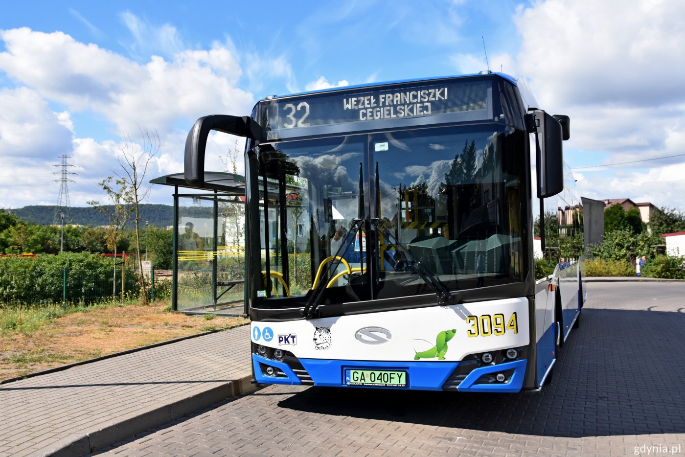 Stojący na przystanku trolejbus. Pojazd nowoczesny, standardowej długości w biało-niebieskich barwach. Trolejbus widziany od frontu. Na wyświetlaczu nr linii 32 i napis 