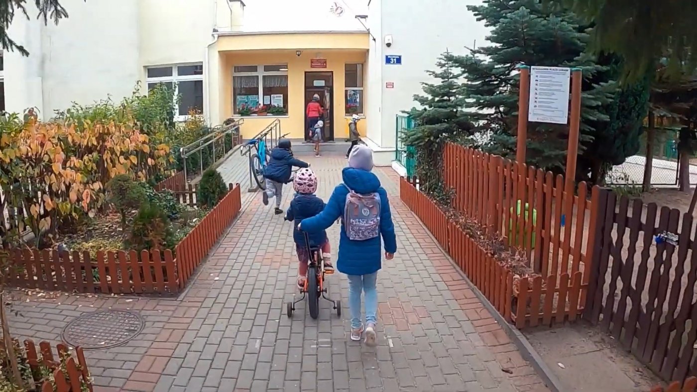 Ścieżka prowadząca do wejścia do szkoły lub przedszkola. Na ścieżce dzieci idące w kierunku wejścia. Niektóre z nich biegną, niektóre jadą rowerem czy hulajnogą.