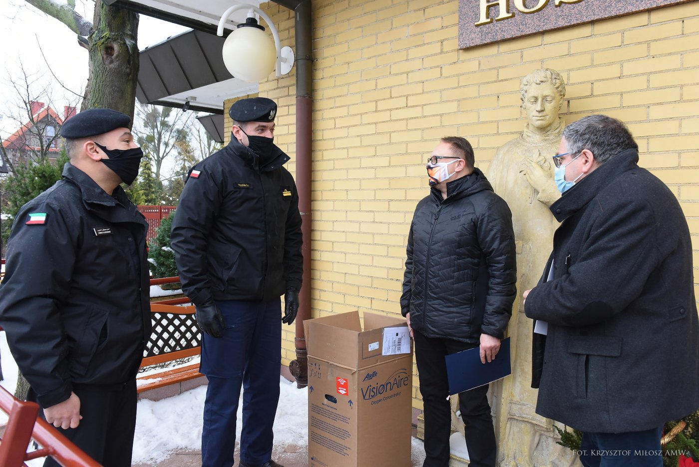 Cztery osoby w ciemnych kurtkach i maseczkach ochronnych rozmawiają przed wejściem do gdyńskiego Hospicjium im. św. Wawrzyńca. Pomiędzy nimi karton z napisem 