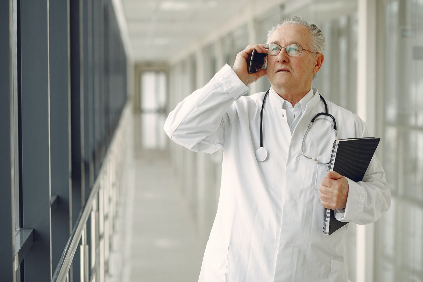 Lekarz ubrany w biały kitel stoi na pustym, szpitalnym korytarzu i rozmawia przez telefon. Na szyi ma stetoskop, a w lewej ręce trzyma zeszyt.