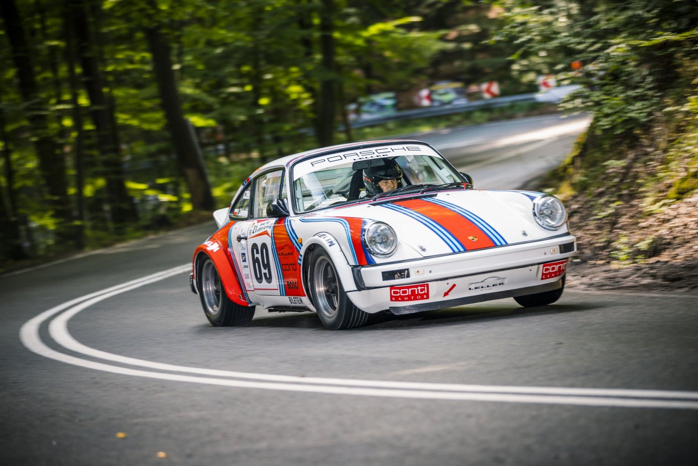 Asfaltowa droga w lesie. Wyraźnie jadąca pod górę z widocznym zakrętem. Na zdjęciu biało-czerwony samochód sportowy marki Porsche.
