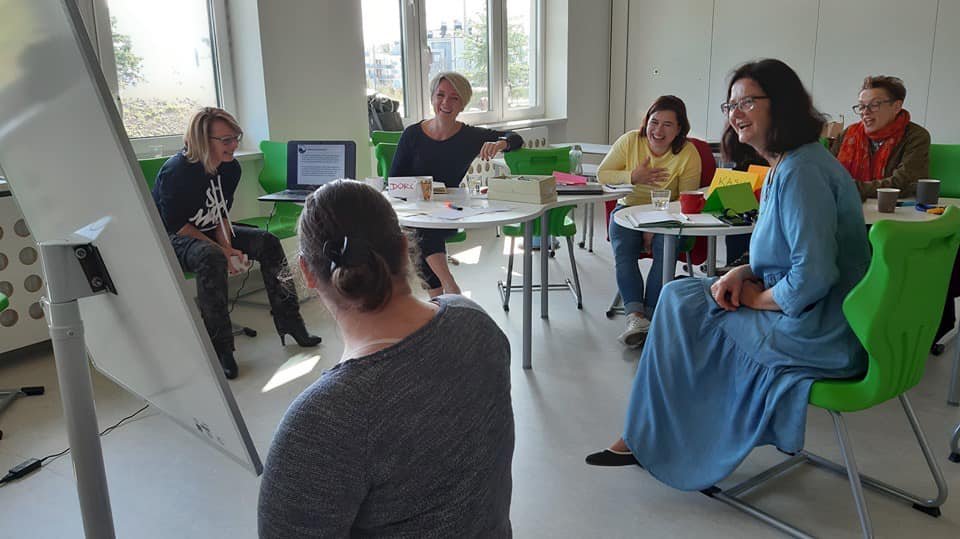 Nauczyciele siedzą w sali lekcyjnej na zielonych krzesłach wokół białego okrągłego stołu i rozmawiają podczas szkolenia