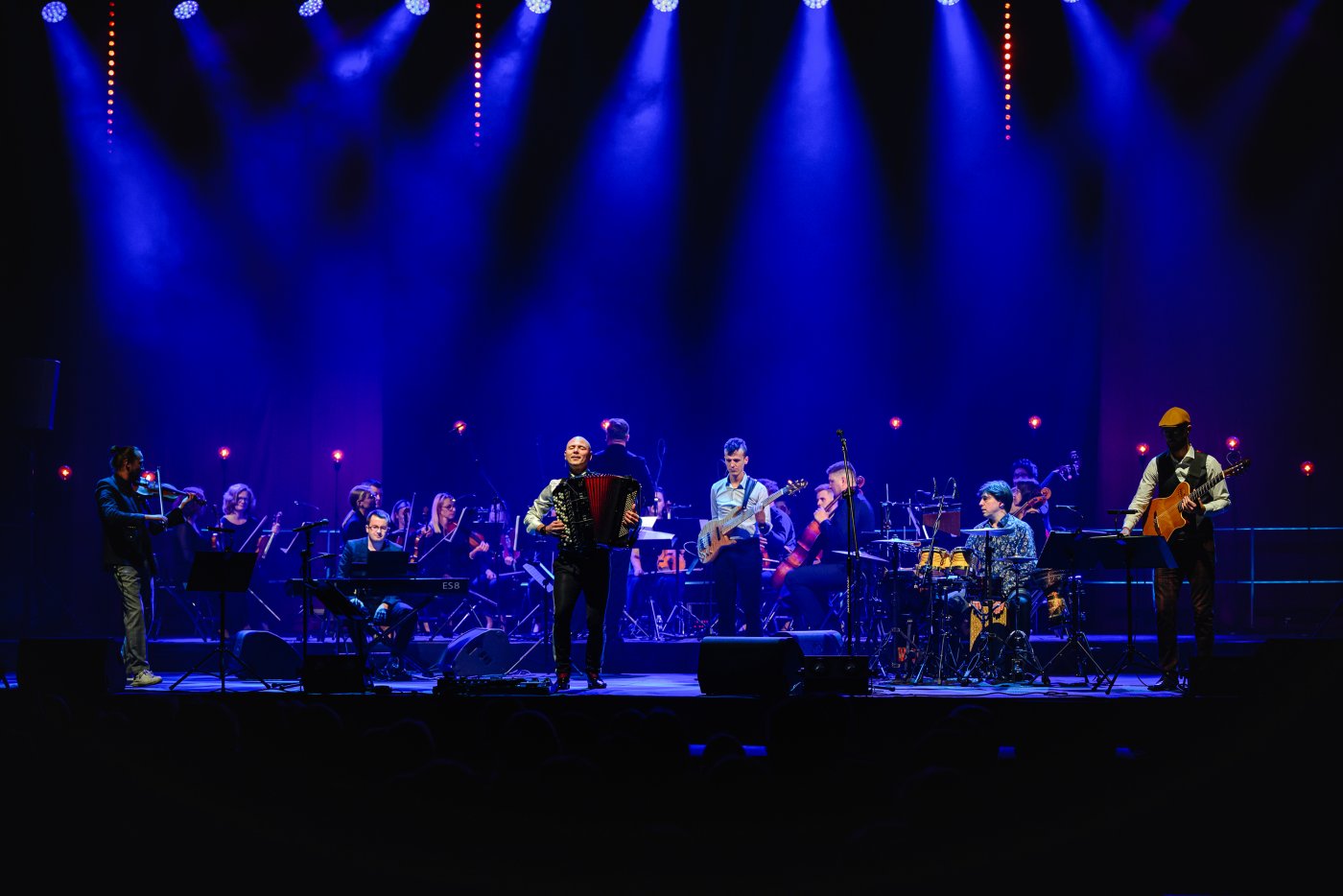 Oświetlona niebieskimi reflektorami scena. Na scenie zespół muzyczny, na pierwszym planie wyraźnie widoczny mężczyzna z akordeonem.