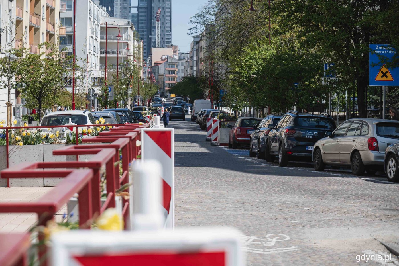 Ulica Starowiejska, widoczne samochody na miejscach parkingowych, budynki, czerwona poręcz ogródka gastronomicznego, słoneczny dzień