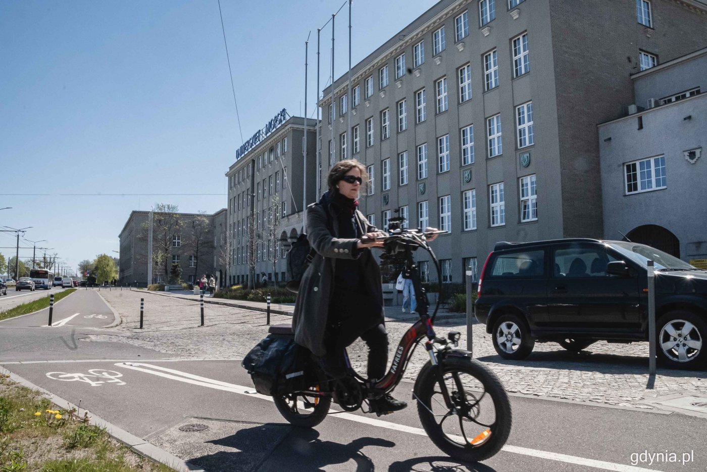 Budynek Uniwersytetu Morskiego, plac przed budynkiem, zaparkowany samochód, na pierwszym planie przejeżdża kobieta na czarnym rowerze