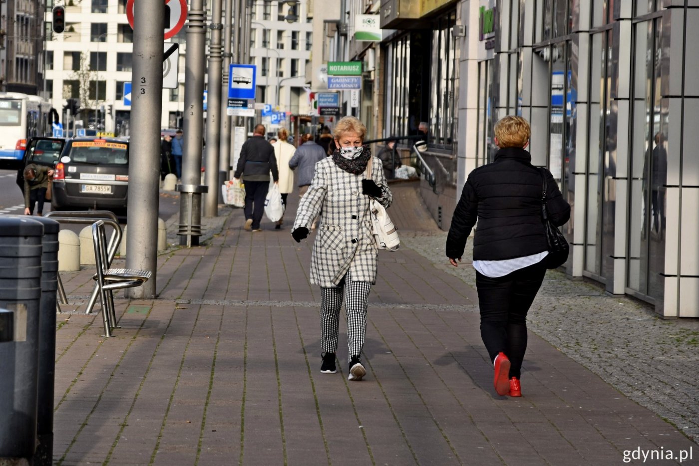 Plac Kaszubski w Gdyni. Ujęcie na chodnik, na którym widać przechodniów. Na pierwszym planie kobieta w maseczce zakrywającej usta i nos. W tle inni przechodnie oraz zabudowa placu Kaszubskiego w Gdyni.