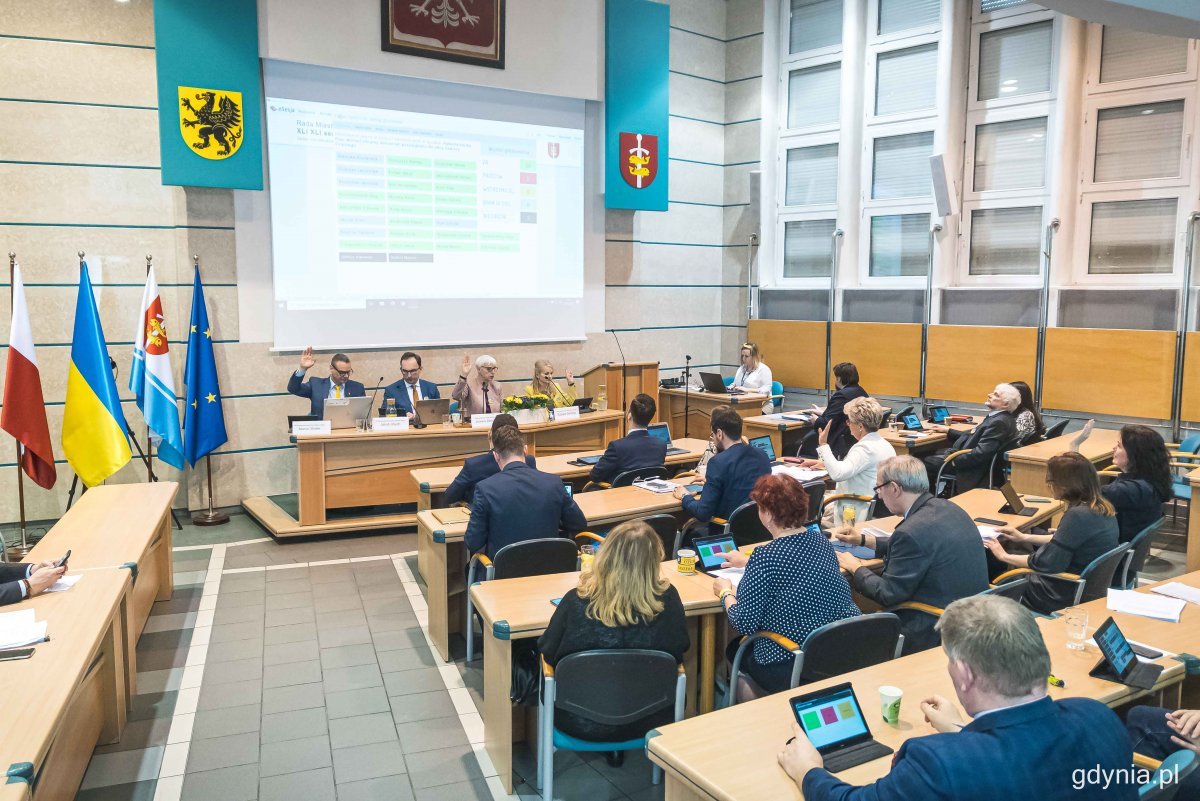 Sala 105 w Urzędzie Miasta Gdyni. Sesja Rady Miasta. Radni siedzą przy stołach, podnoszą ręce głosując. Na ekranie informacje o głosowaniu