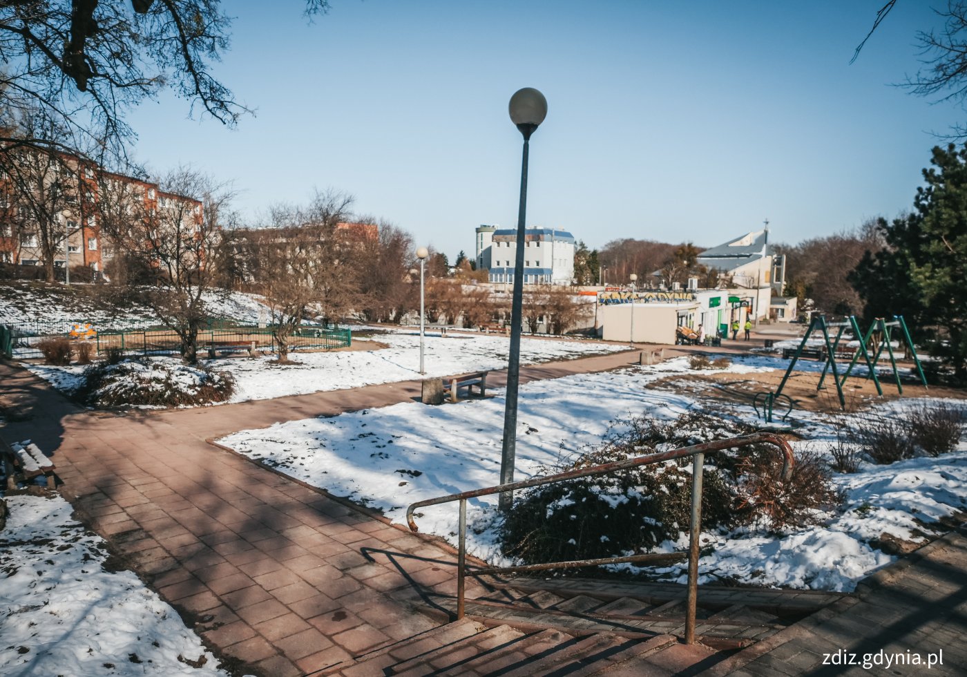 na zdjęciu widoczny skwer ze sprzętami służącymi do zabawy, na zdjęciu widoczne są schody z barierką, huśtawki, ławki oraz ogrodzony plac, zieleń