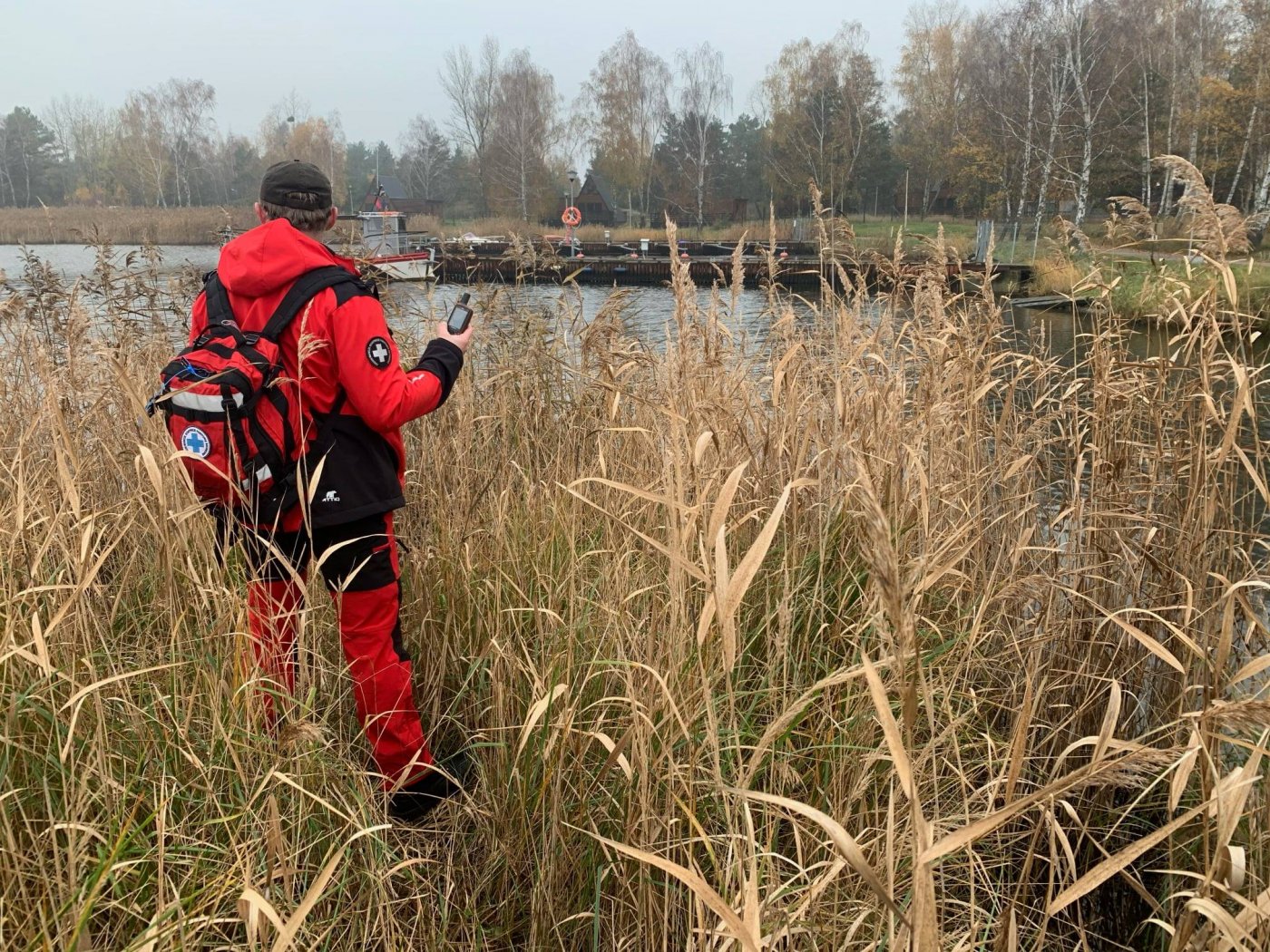 Na pierwszym planie ratownik medyczny (po lewej stronie zdjęcia) ubrany w czerwony uniform oznaczony. W ręku trzyma walkie-talkie. Stoi w zaroślach typowych dla terenów leśnych i jeziornych. W drugim planie mały staw w środku lasu.