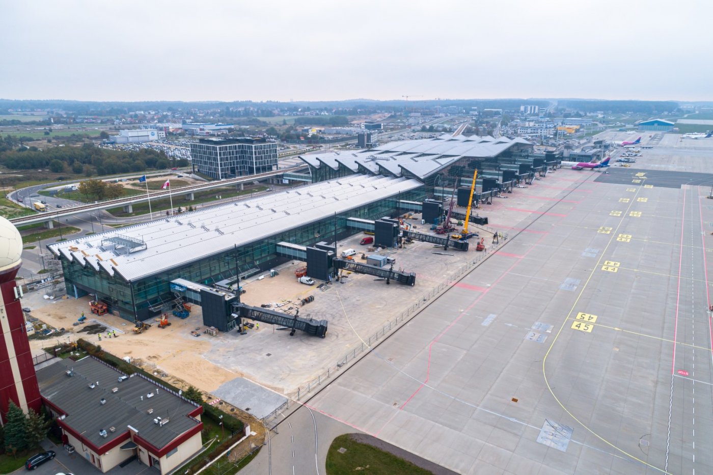 Zdjęcie lotnicze. Widoczny Port Lotniczy w Gdańsku. Rozbudowany budynek terminala, dźwigi montują rękawy pasażerskie. Płyta lotniska