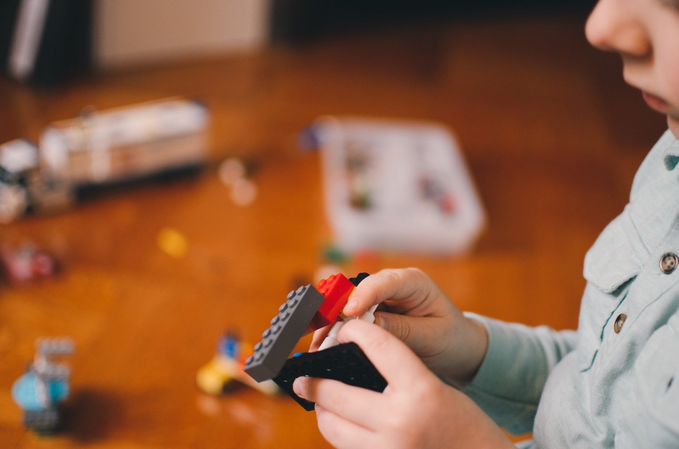 Na zdjęciu widać rozrzucone kolorowe klocki LEGO, z których ręka dziecka buduje robota na tle brązowej podłogi.