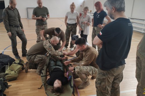 Sala gimnastyczna, grupa ludzi przechodzi szkolenie z pierwszej pomocy, cztery osoby w mundurach kucają przy osobie na noszach, kilka osób obserwuje ćwiczenia, dookoła militarne plecaki